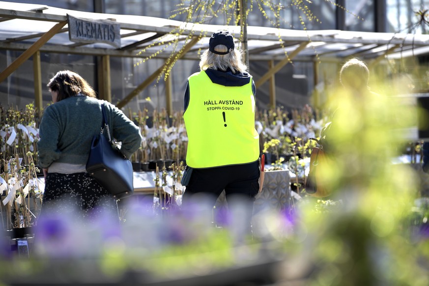 epa08375387 An employee wears a vest with a text reading &quot;Keep distance. Stop Covid-19&quot; as customers look at plants at the Slottstradgarden Ulriksdal garden centre situated in the Ulriksdal  ...