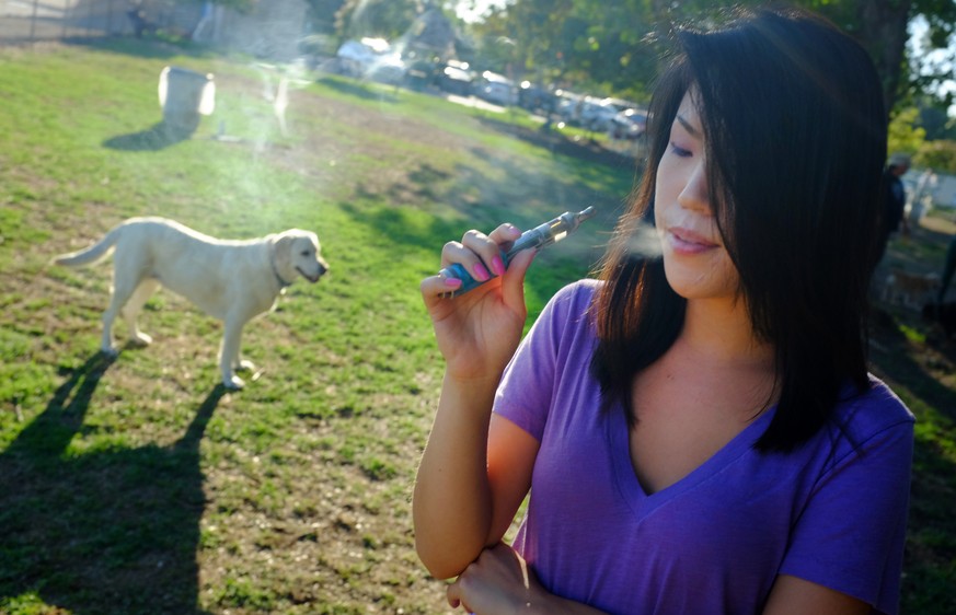 In this photo taken on Tuesday, July 1, 2014, Christine Choi takes a puff on her e-cigarette at the Sepulveda Basin Dog Park in Los Angeles, as she accompanies her dog, not pictured. Recent surges in  ...