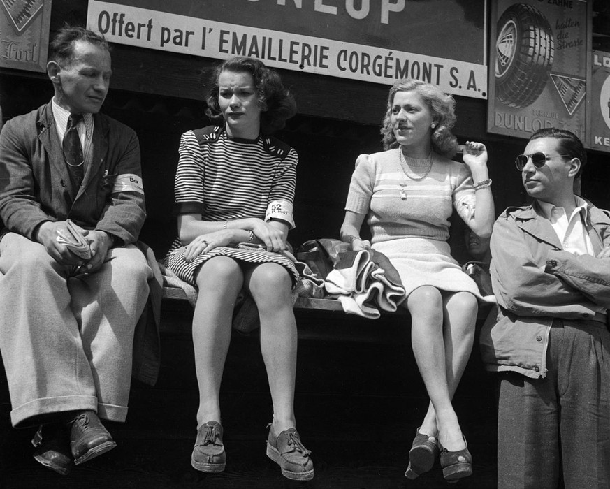 Zuschauerinnen und Zuschauer lassen ihre Beine unter einem Emaille-Schild der Firma Dunlop baumeln, aufgenommen am Grossen Preis der Schweiz in Bremgarten im Juni 1947. (KEYSTONE/Photopress-Archiv/Str ...