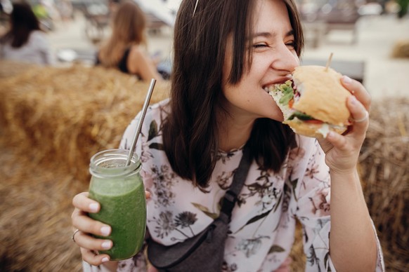 Stilvolles hipster Mädchen in einer Sonnenbrille, das köstliche veganische Burger isst und Smoothie in Glaskeramik in Händen hält. Happy boho Frau beissen Burger mit Drink in der Sommerstraße