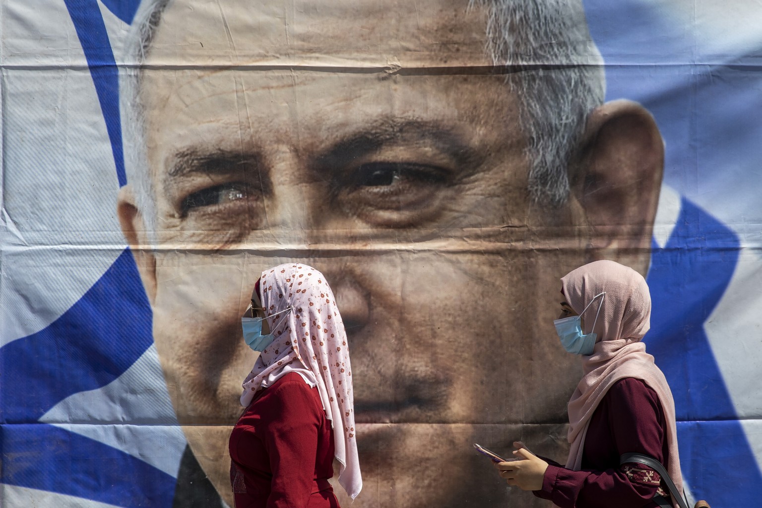Muslim women wear face masks to help protect themselves from the coronavirus, walk past a poster hung by supporters of Prime Minister Benjamin Netanyahu outside the district court in Jerusalem, Sunday ...