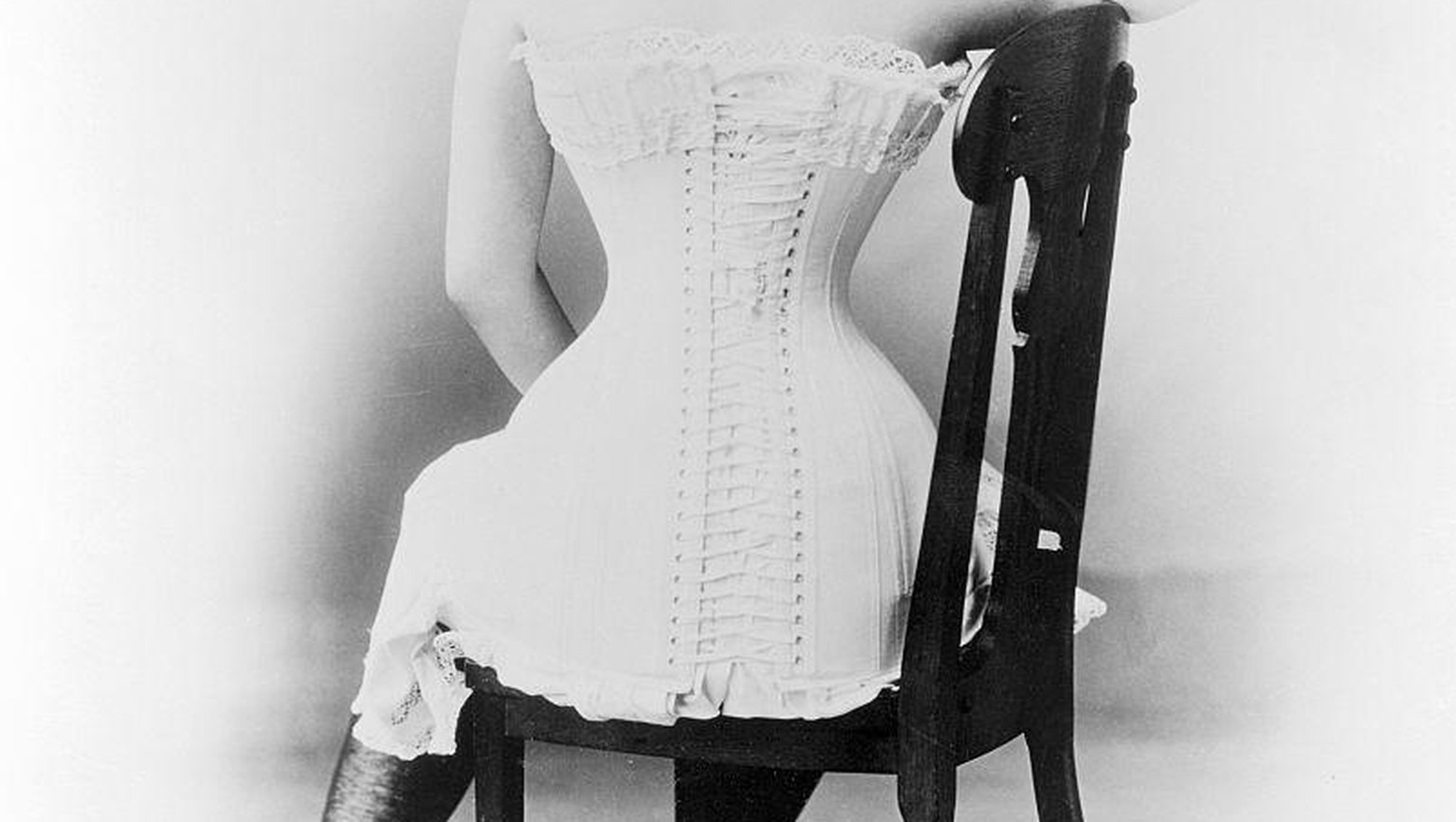 ORIGINAL CAPTION READS: Young lady in victorian corset. Back view seated in chair. Undated photograph.