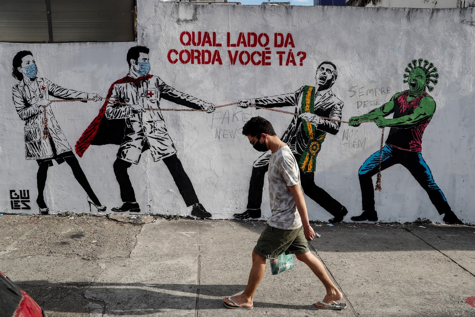 epa08504708 A man passes in front of a graffiti that represents Brazilian President Jair Bolsonaro and a figure that represents the novel coronavirus COVID-19 pulling a rope against health workers wit ...