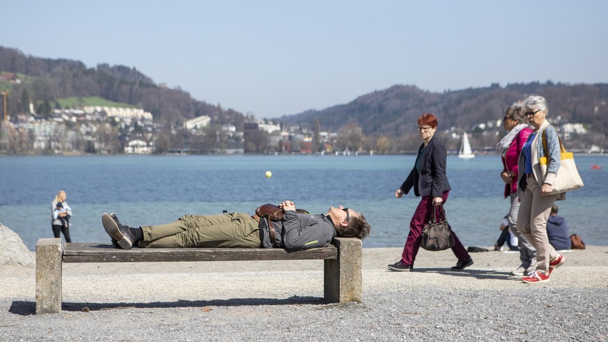 Ein Mann liegt in der Sonne auf einer Parkbank neben Passanten in der Ufschoetti in Luzern am Samstag, 7.April 2018, in Luzern. (KEYSTONE/Patrick Huerlimann)