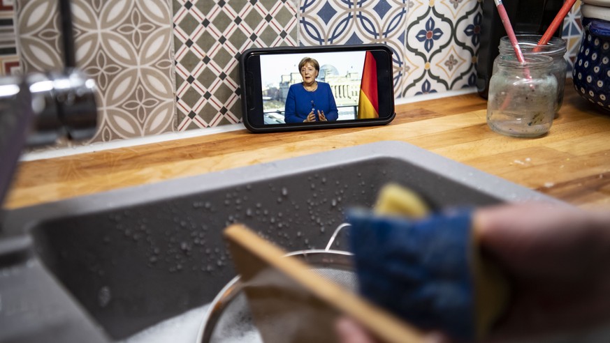 epa08304857 The illustration shows German Chancellor Angela Merkel, during her televised address t?o the nation on the coronavirus spread, on a smartphone while a woman is cleaning dishes in Munich, B ...