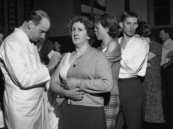 Bei einer freiwilligen Impfaktion der Bevoelkerung von Schaffhausen am 8. Januar 1962 impft ein Arzt eine Frau in den entbloessten Oberarm, waehrend die anderen Leute in einer Reihe warten. Da ein jun ...