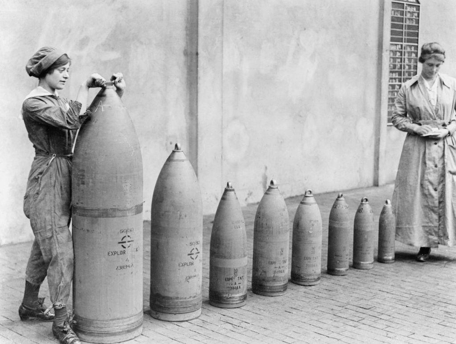 Women At Work During The First World War: Munitions Production, Chilwell, Nottinghamshire, England, UK, c. 1917, Two women munitions workers stand beside examples of the shells produced at National Sh ...