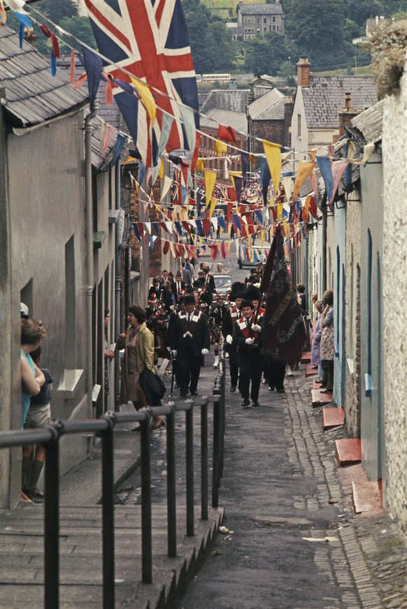 View of an annual Apprentice Boys&#039; parade, with Protestant pipe band members marching and playing along a street decked with bunting and union flags, to celebrate the lifting of the siege of the  ...