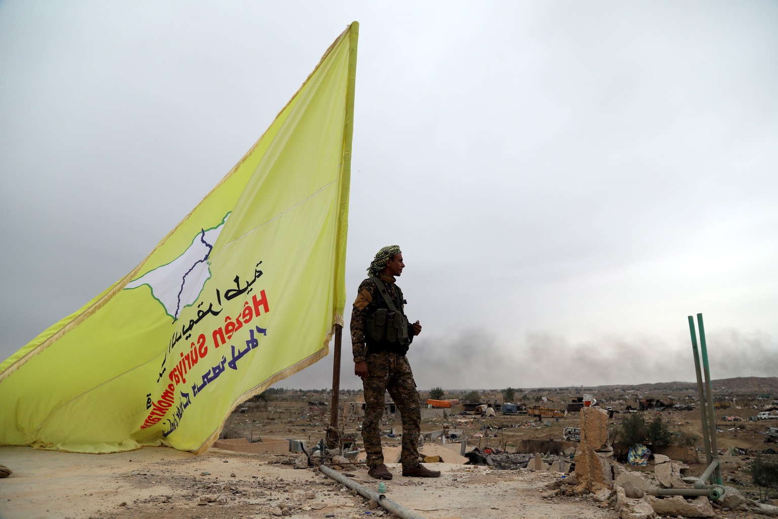 epaselect epa07461235 A Fighter of the Syrian Democratic Forces (SDF) stands guard next to the yellow flag of US-backed Syrian Democratic Forces (SDF) atop a building in the village of Baghuz, Syria,  ...