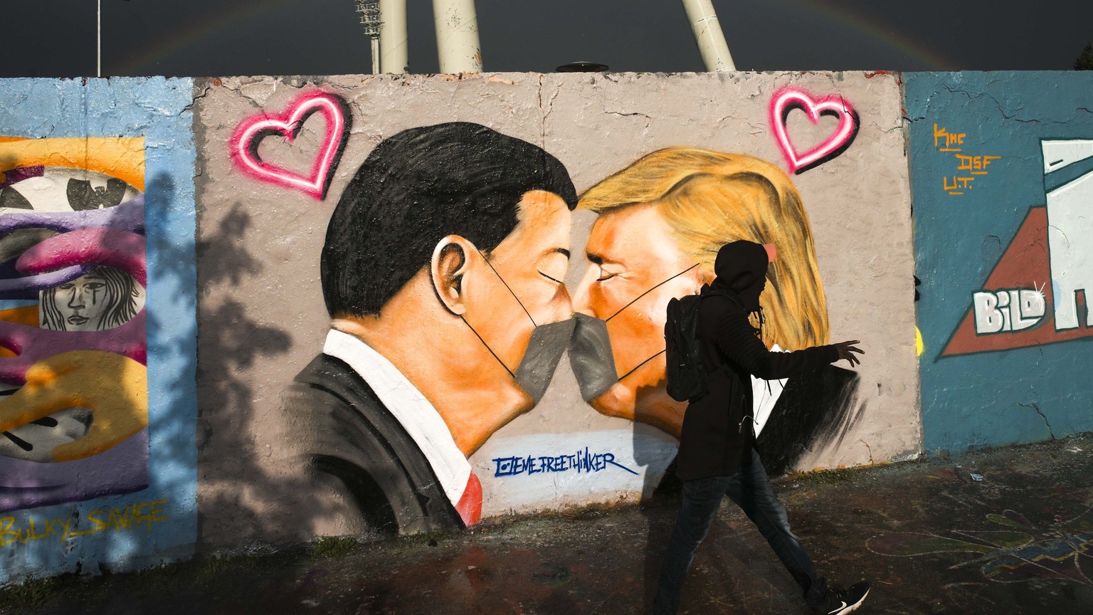 A man walks in front of graffiti depicting US President Trump, right, and China&#039;s President Xi Jinping kissing each other with face masks, displayed at a wall in the public park Mauerpark in Berl ...