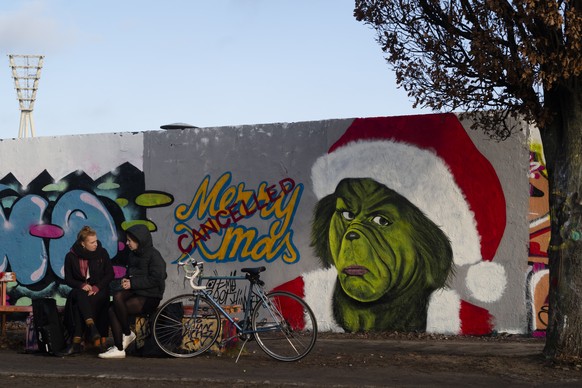 Two women sit on a bench in front of a graffiti by artist &#039;EME Freethinker&#039; depicting the movie character &#039;The Grinch&#039; and the slogan &#039;Merry Christmas - Cancelled&#039; at the ...