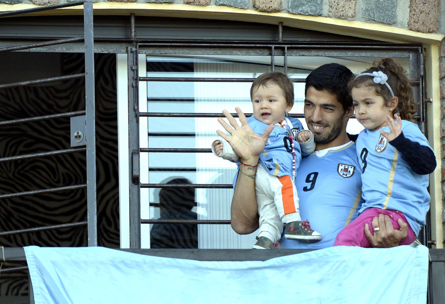 Vor dem Spiel gegen Kolumbien noch zuversichtlich: Luis Suarez winkt vom Balkon seines Hauses in Montevideo, gemeinsam mit seinen Kindern Benjamin und Delfina.